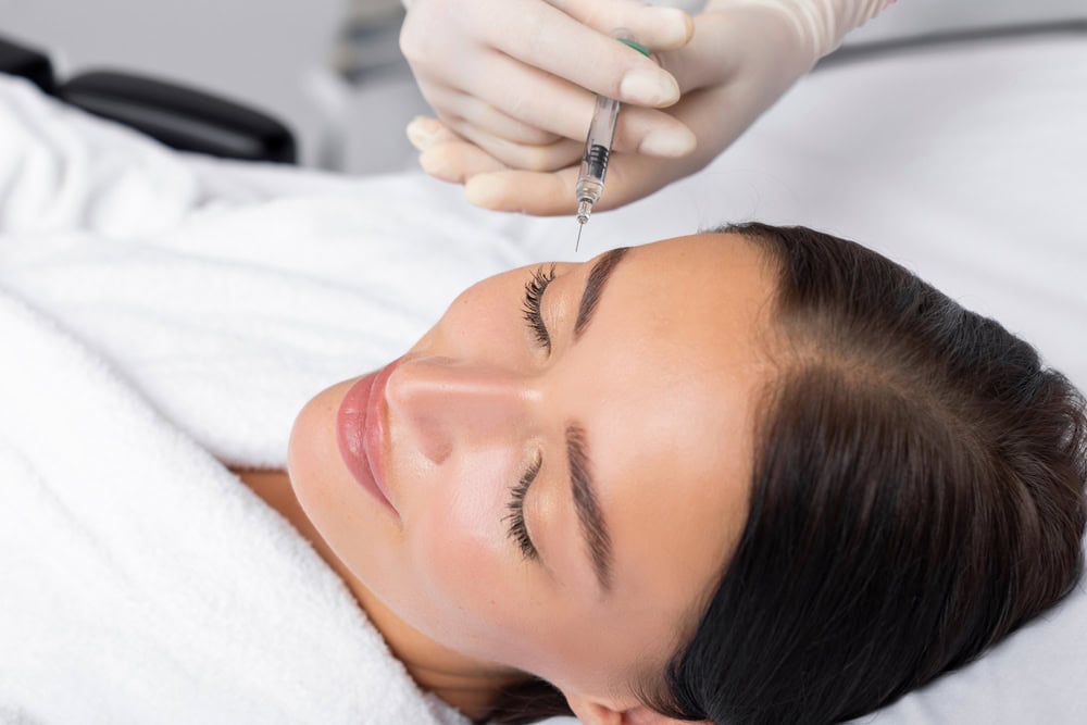 A woman in a white robe receives a dermal filler injection near her eyebrow from a gloved hand.